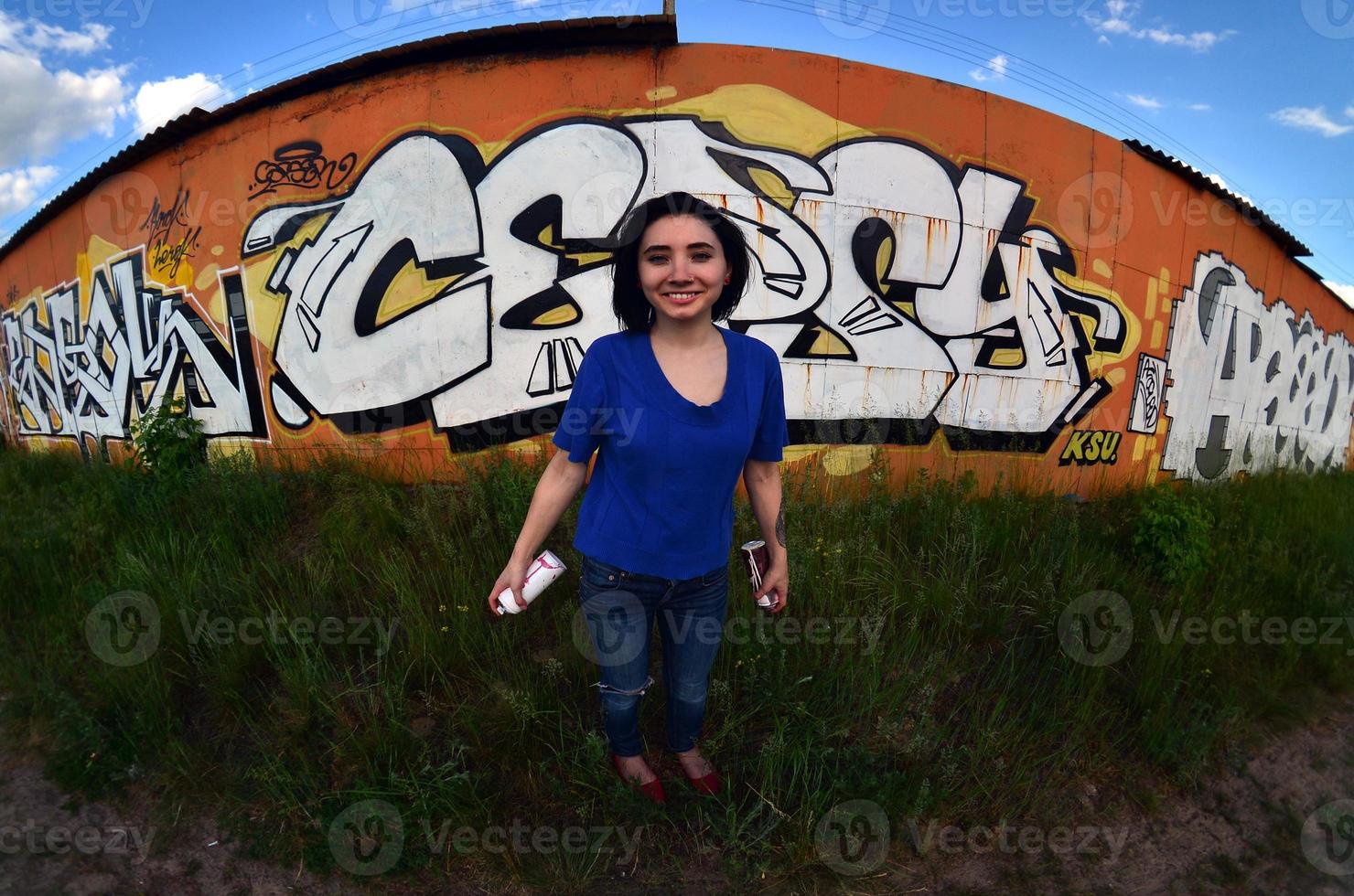 portret van een emotioneel jong meisje met zwart haar- en piercings. een wijde hoek foto van een meisje met aërosol verf blikjes in de handen Aan een graffiti muur achtergrond. een modern portret van een vissenoog lens