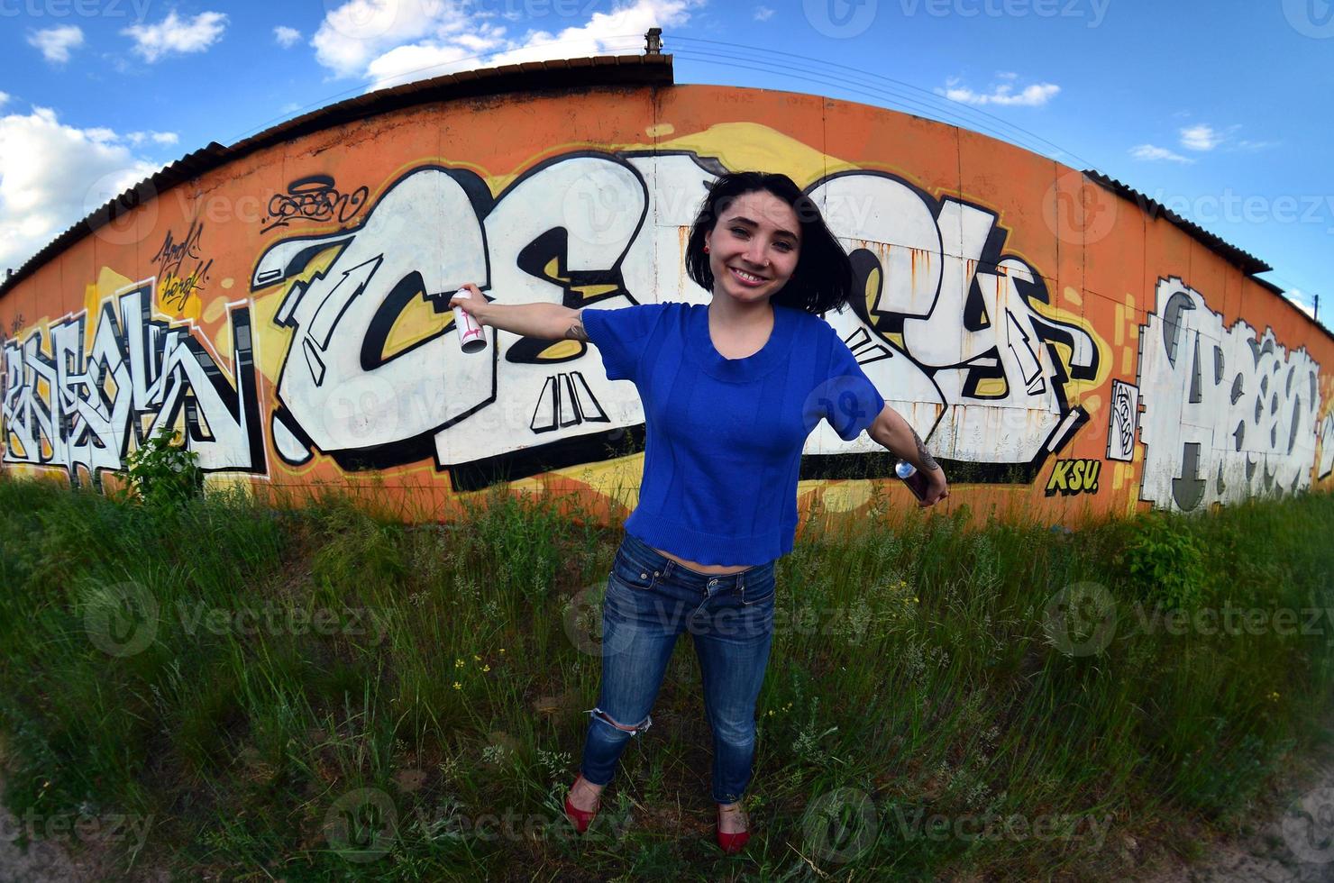 portret van een emotioneel jong meisje met zwart haar- en piercings. een wijde hoek foto van een meisje met aërosol verf blikjes in de handen Aan een graffiti muur achtergrond. een modern portret van een vissenoog lens