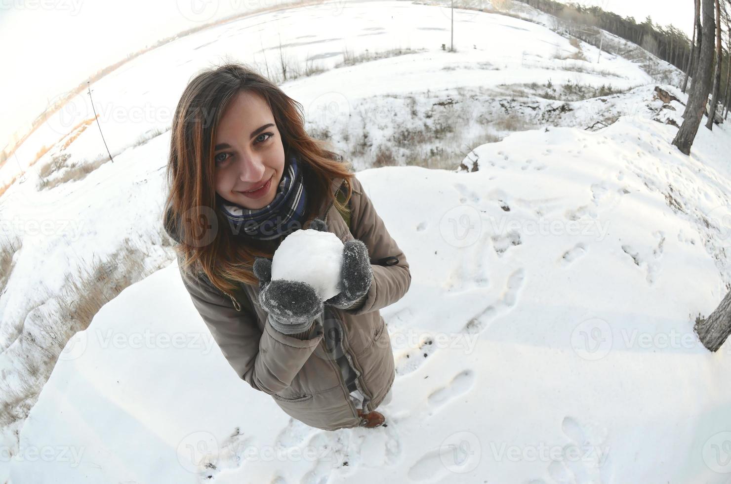 een jong en blij Kaukasisch meisje in een bruin jas houdt een sneeuwbal in voorkant van een horizon lijn tussen de lucht en een bevroren meer in winter. vissenoog foto