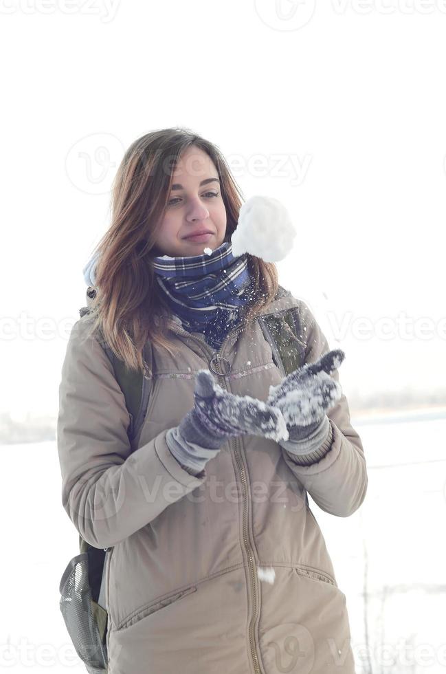 een jong en blij Kaukasisch meisje in een bruin jas houdt een sneeuwbal in de achtergrond van een horizon lijn tussen de lucht en een bevroren meer in winter foto