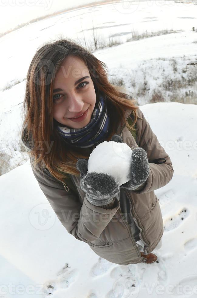 een jong en blij Kaukasisch meisje in een bruin jas houdt een sneeuwbal in voorkant van een horizon lijn tussen de lucht en een bevroren meer in winter. vissenoog foto