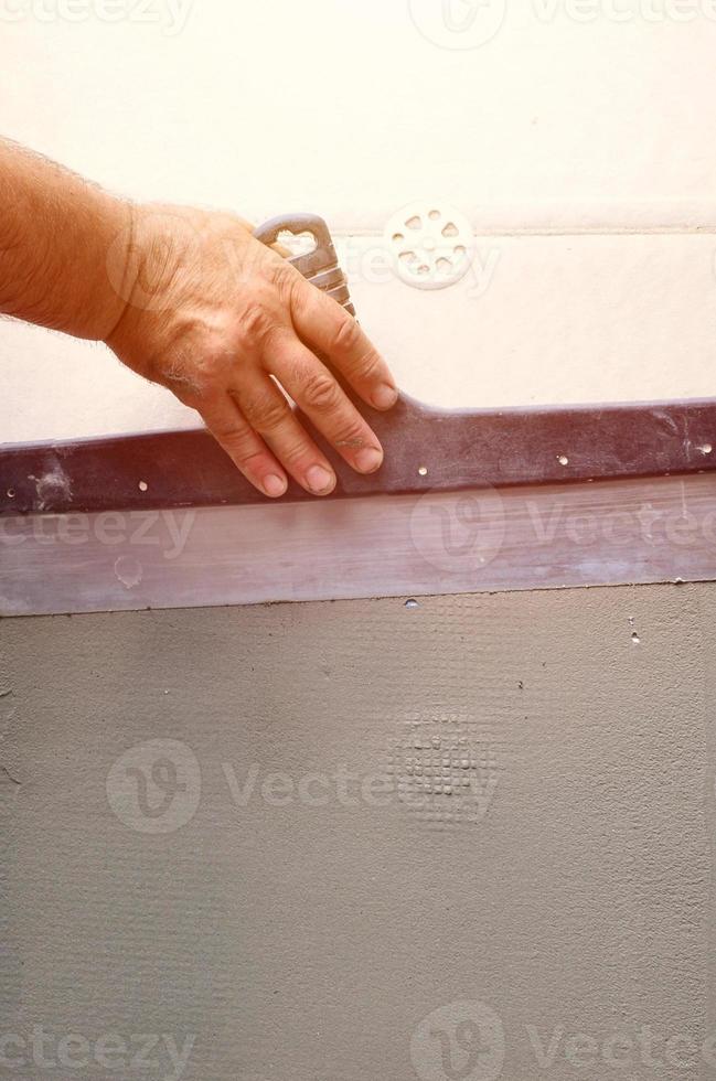 handen van een oud handleiding arbeider met muur bepleistering gereedschap renoveren huis. stukadoor renoveren buitenshuis muren en hoeken met spatel en gips. muur isolatie. bouw afwerking werken foto