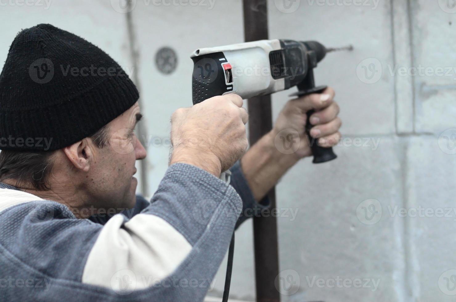 een ouderen werkman boren een gat in een piepschuim muur voor de volgend installatie van een plastic versterken deuvel. creëren gaten in de muur met een oefening. opwarming van de gebouw facade foto