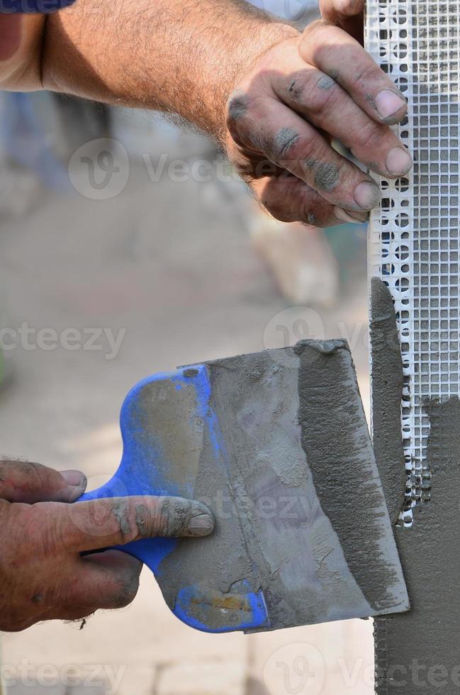 handen van een oud handleiding arbeider met muur bepleistering gereedschap renoveren huis. stukadoor renoveren buitenshuis muren en hoeken met spatel en gips. muur isolatie. bouw afwerking werken foto