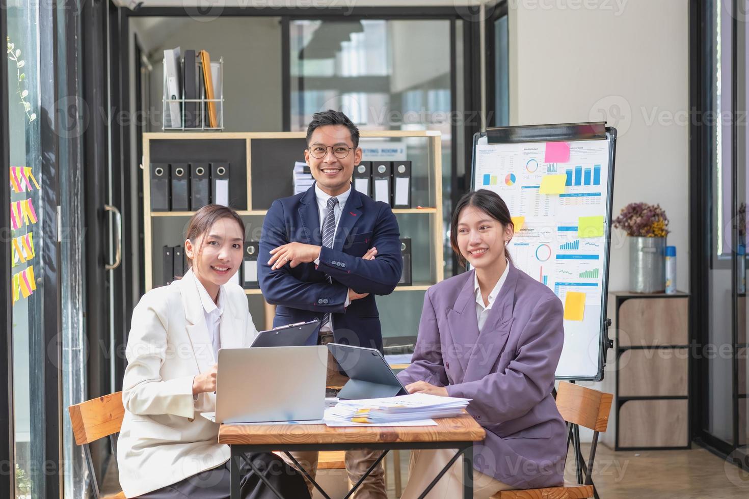 team bedrijf drie glimlachen en gelukkig Aziatisch mannen en Dames team gebruik makend van tablets en laptops brainstorm en verhaal statistieken Aan een zakelijke tabel bord binnen de bedrijf. collega's concept. foto