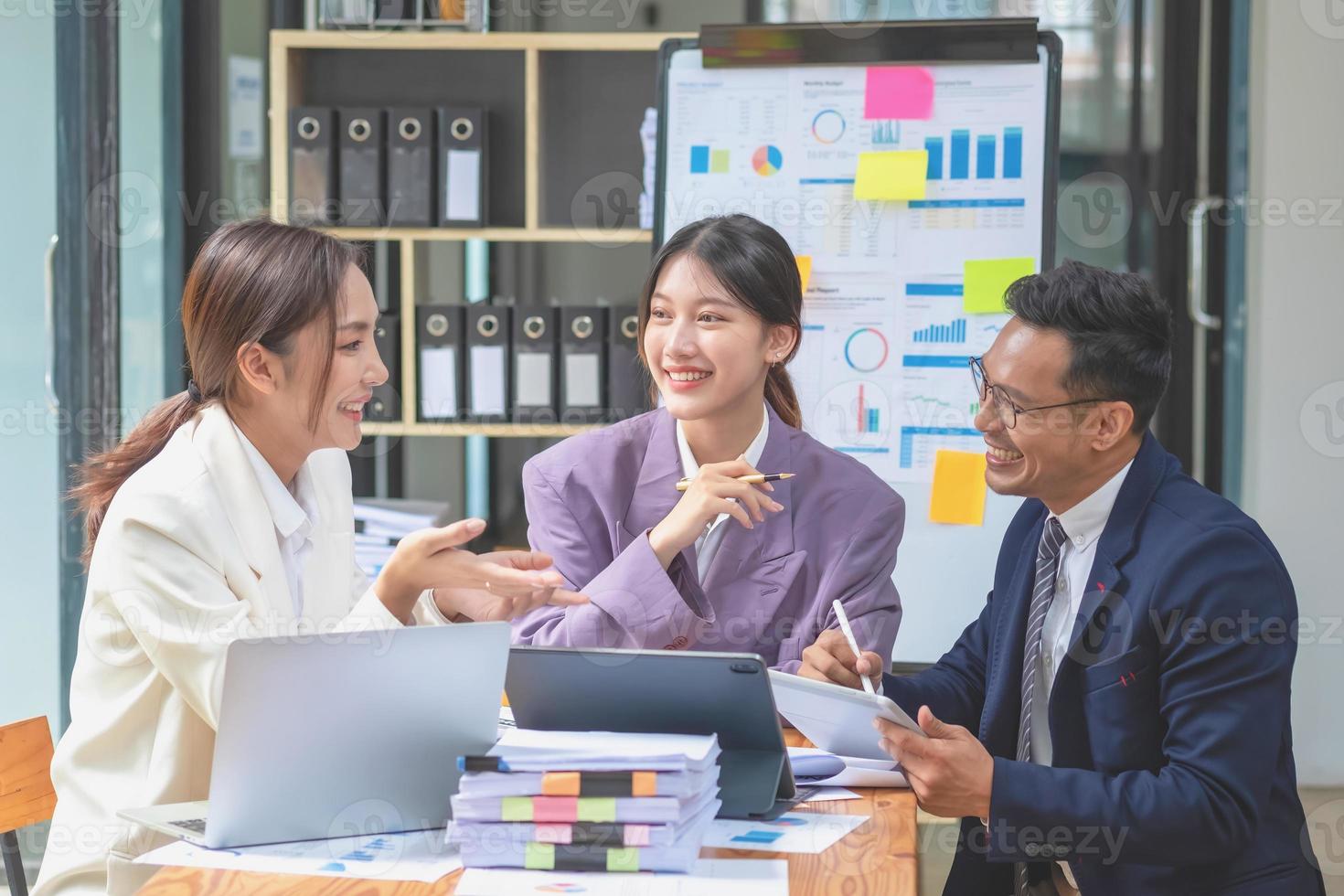 team bedrijf drie glimlachen en gelukkig Aziatisch mannen en Dames team gebruik makend van tablets en laptops brainstorm en verhaal statistieken Aan een zakelijke tabel bord binnen de bedrijf. collega's concept. foto