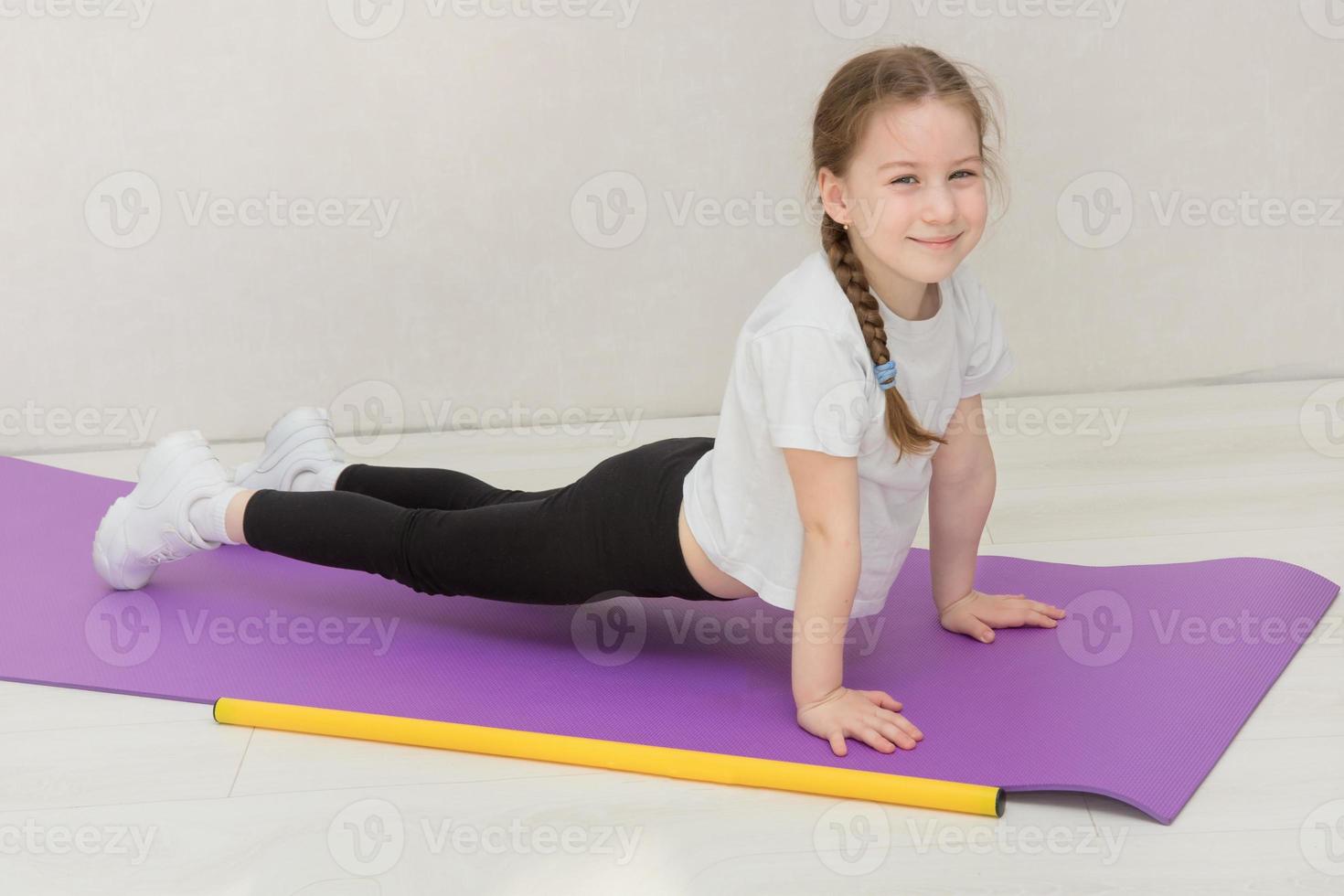 schattig weinig meisje staat in een plank Aan een mat, een gymnastiek- stok leugens dichtbij, de kind glimlacht foto