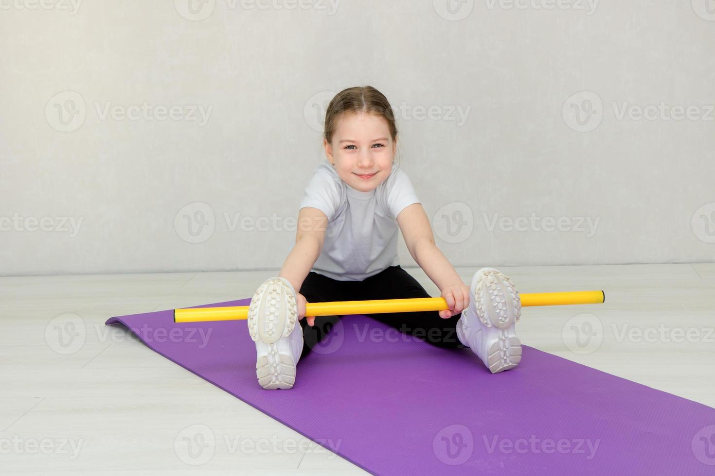 schattig weinig meisje zittend Aan een mat en aan het doen opdrachten met een gymnastiek- stok, kinderen geschiktheid foto