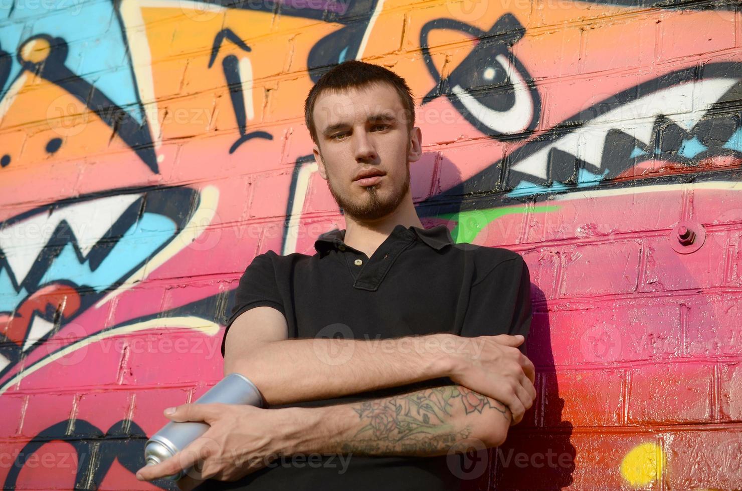 jong Kaukasisch graffiti artiest in zwart t-shirt met zilver aërosol verstuiven kan in de buurt kleurrijk graffiti in roze tonen Aan steen muur. straat kunst en hedendaags schilderij werkwijze foto