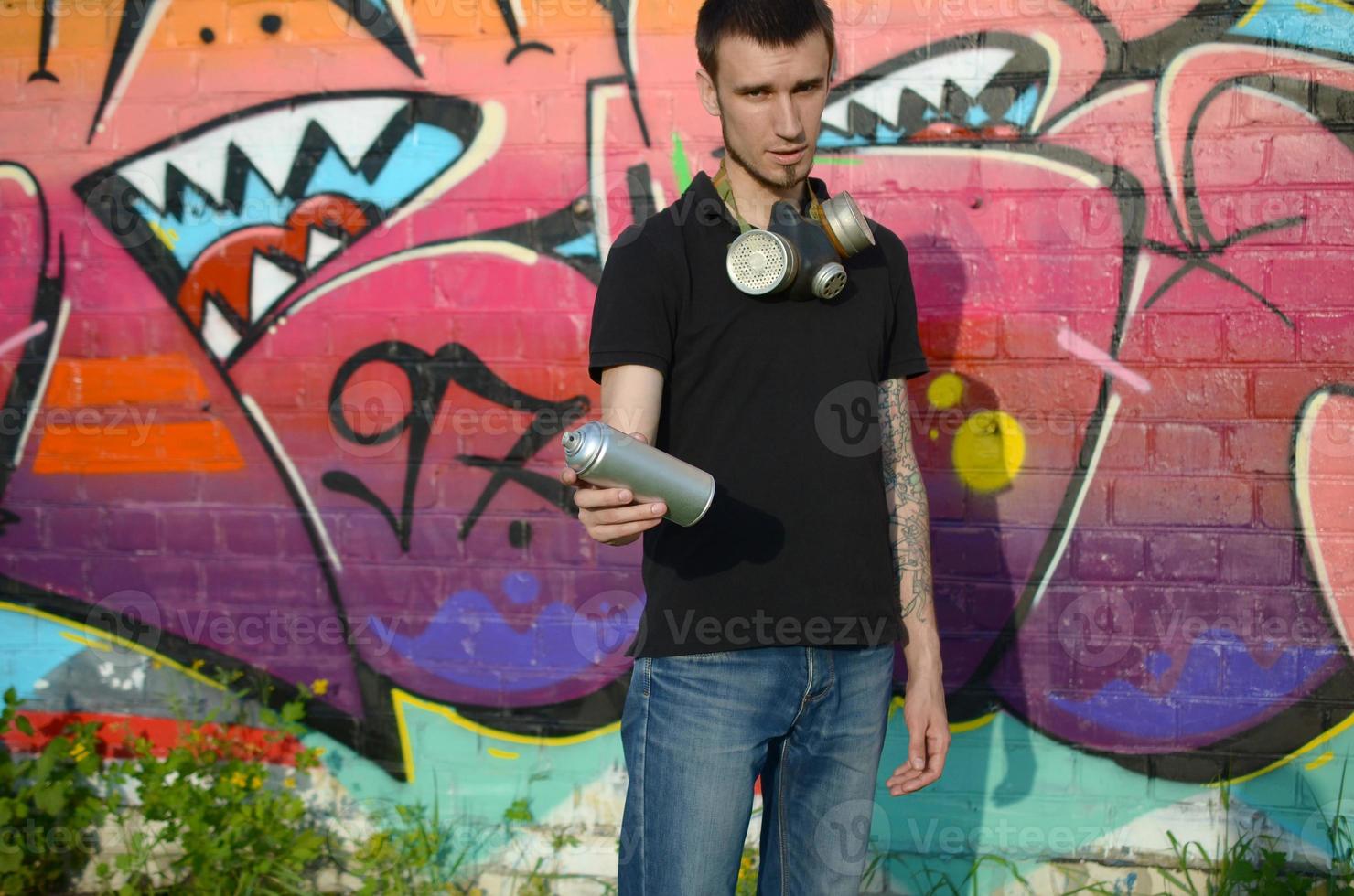 jong Kaukasisch graffiti artiest in zwart t-shirt met zilver aërosol verstuiven kan in de buurt kleurrijk graffiti in roze tonen Aan steen muur. straat kunst en hedendaags schilderij werkwijze foto