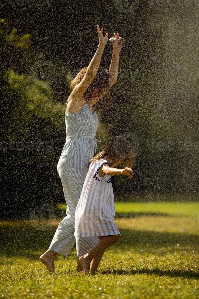 schattig weinig meisje hebben pret onder irrigatie sproeier met haar moeder foto