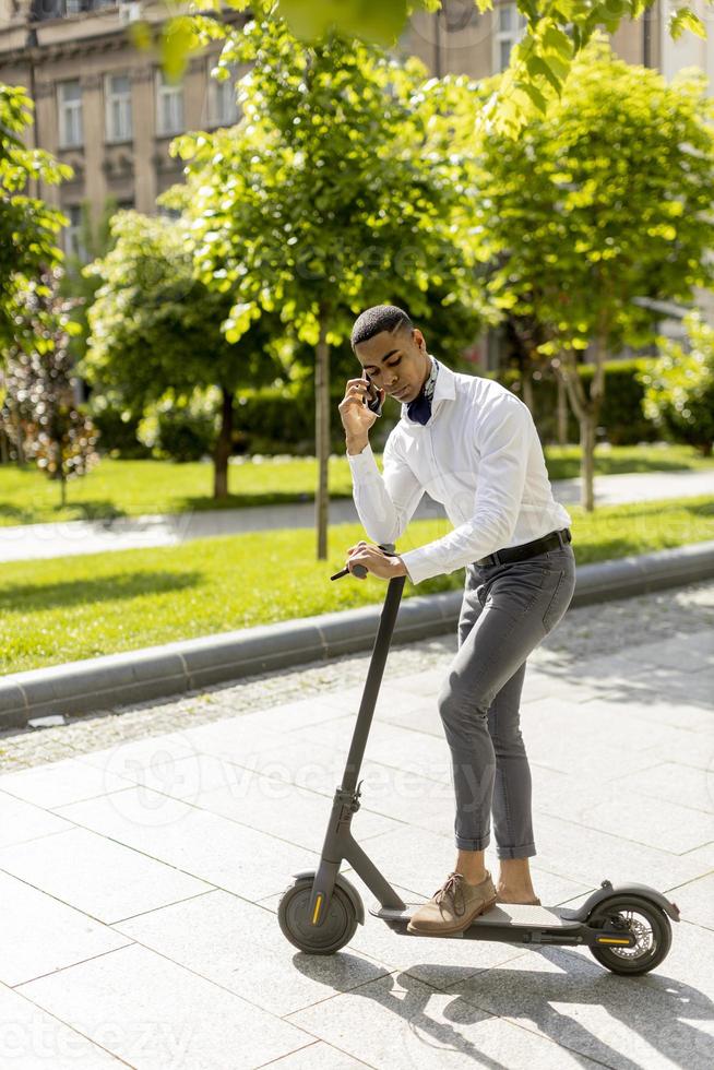 jonge Afro-Amerikaan die mobiele telefoon gebruikt terwijl hij met een elektrische scooter op straat staat foto