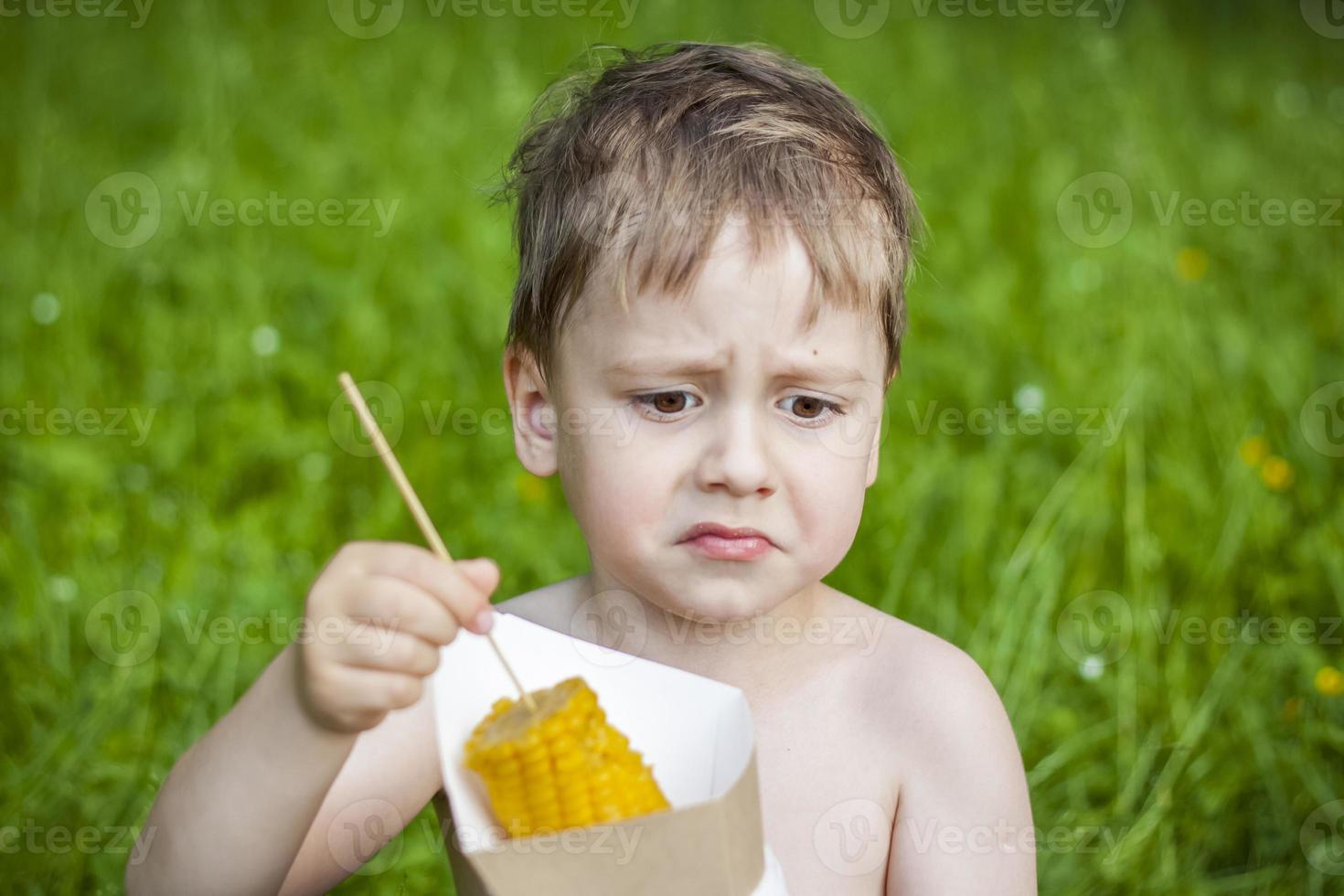 een schattig blond jongen smakelijk eet maïs in de zomer, zittend Aan de bank van de rivier- Aan de sappig gras. grappig gelaats uitdrukking. foto