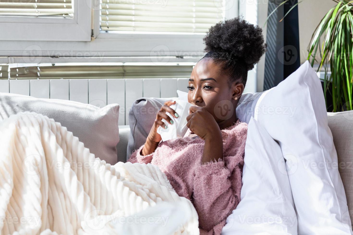 ziek ten einde raad vrouw heeft griep. allergie concept. mooi ziek vrouw heeft rennen neus, wrijft neus- met zakdoek en drinken heet drank. Afrikaanse Amerikaans vrouw niezen in een zakdoek foto