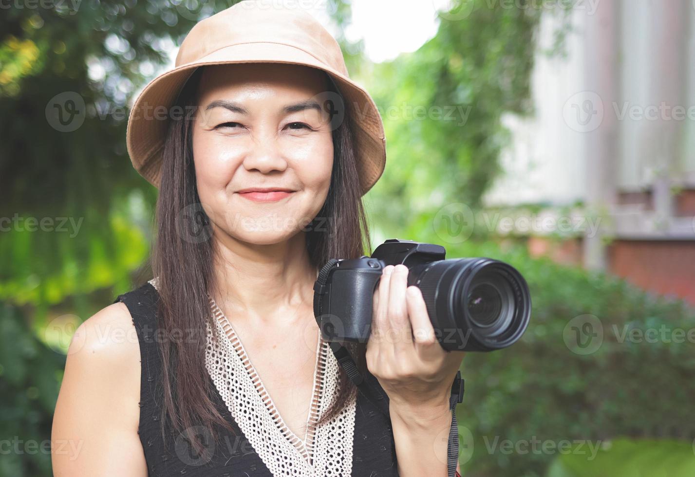 Aziatisch vrouw, vervelend hoed en zwart top mouwloos, staand in de tuin, Holding dslr camera, glimlachen gelukkig. foto