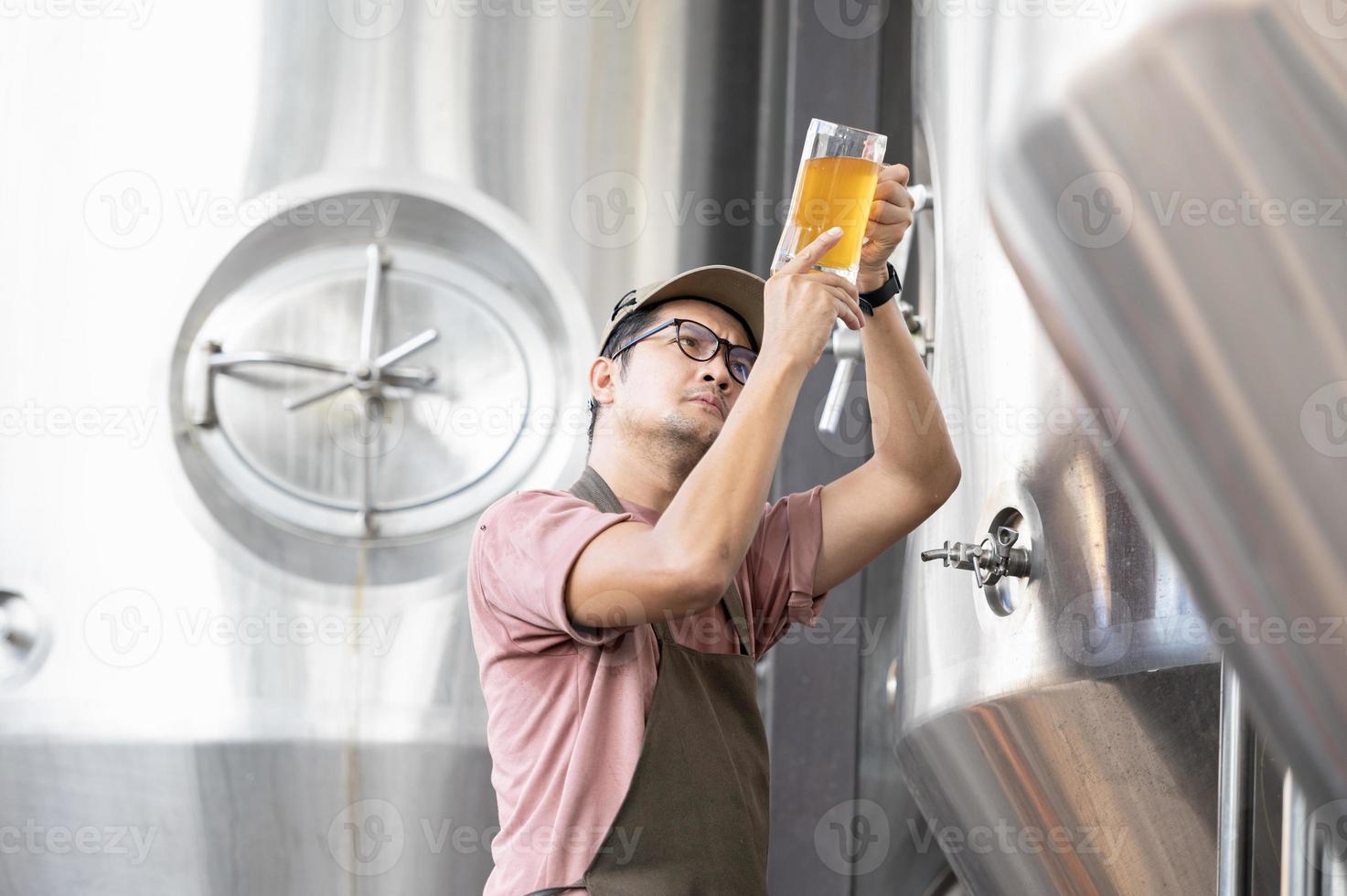 jong Aziatisch arbeider inspecteren brouwerij kwaliteit met een glas van ambacht bier evalueren zichtbaar uiterlijk na voorbereiding terwijl werken in een verwerken ambacht brouwerij. foto