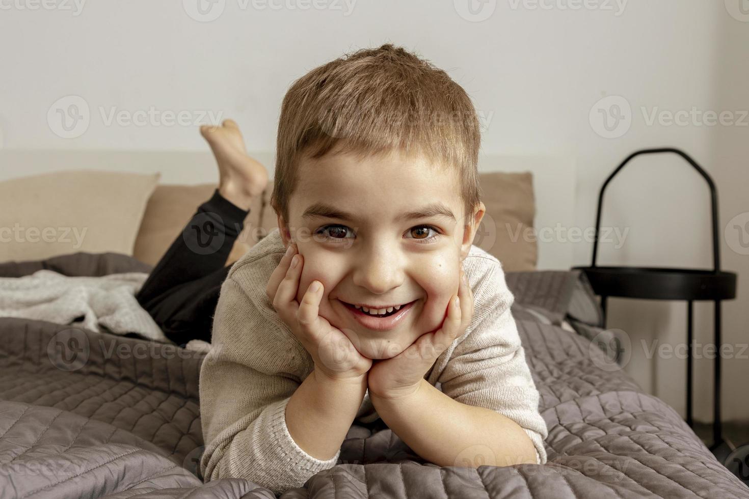 portret van klein, glimlachen Kaukasisch jongen Aan de bed Bij huis. schattig kind ontspannend, resting in slaapkamer. positief emoties. knus en modern interieur. natuurlijk, aarde kleuren. gewoontjes kleren. foto