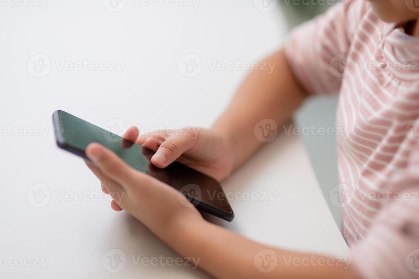 schattig weinig meisje toepassingen smartphone terwijl zittend Bij de sofa in de leven kamer. kind surfing de internet Aan mobiel telefoon, bladert door internet en horloges tekenfilms online Bij huis foto