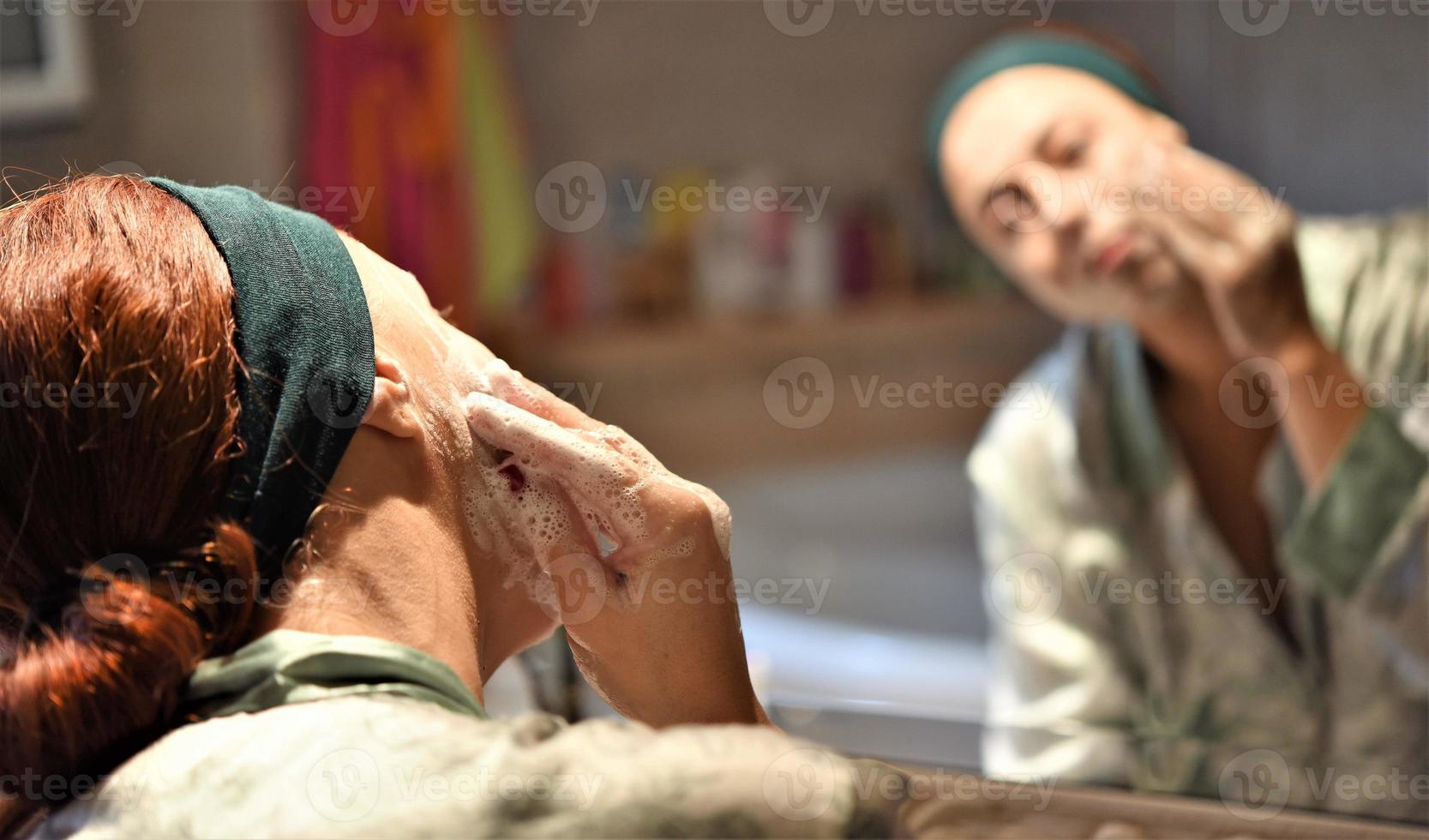 mooi jong vrouw is het wassen haar gezicht en glimlachen terwijl staand in voorkant van de spiegel in de badkamer foto