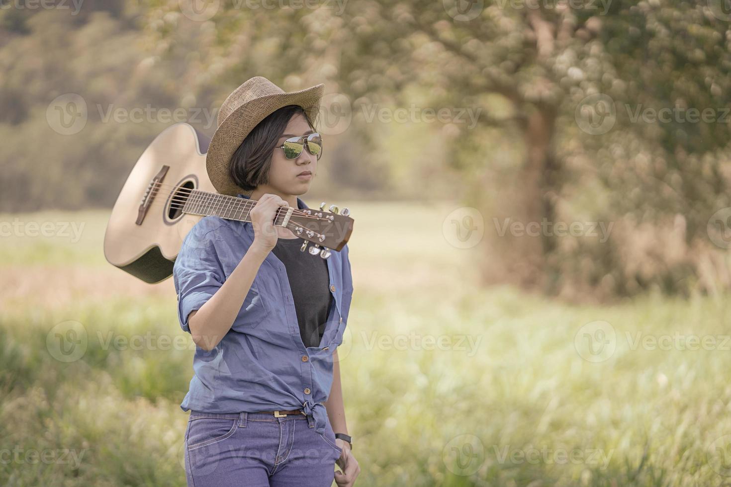 vrouw slijtage hoed en dragen haar gitaar in gras veld- foto