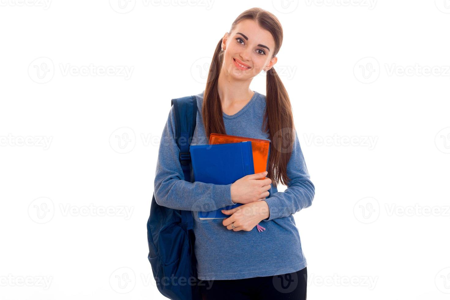 vrolijk jong brunette leerling meisje met blauw rugzak en een veel van boeken in haar handen poseren en op zoek Bij de camera en glimlachen geïsoleerd Aan wit achtergrond foto