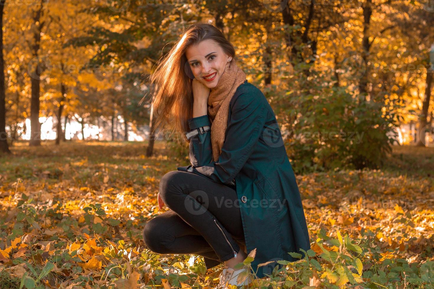 jong schattig meisje in een zwart mantel zit in de park in herfst Aan de gazon en glimlacht foto