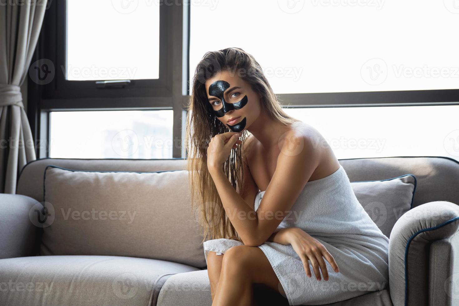 mooi vrouw is ontspannende Bij huis met zwart afpellen masker Aan haar gezicht foto