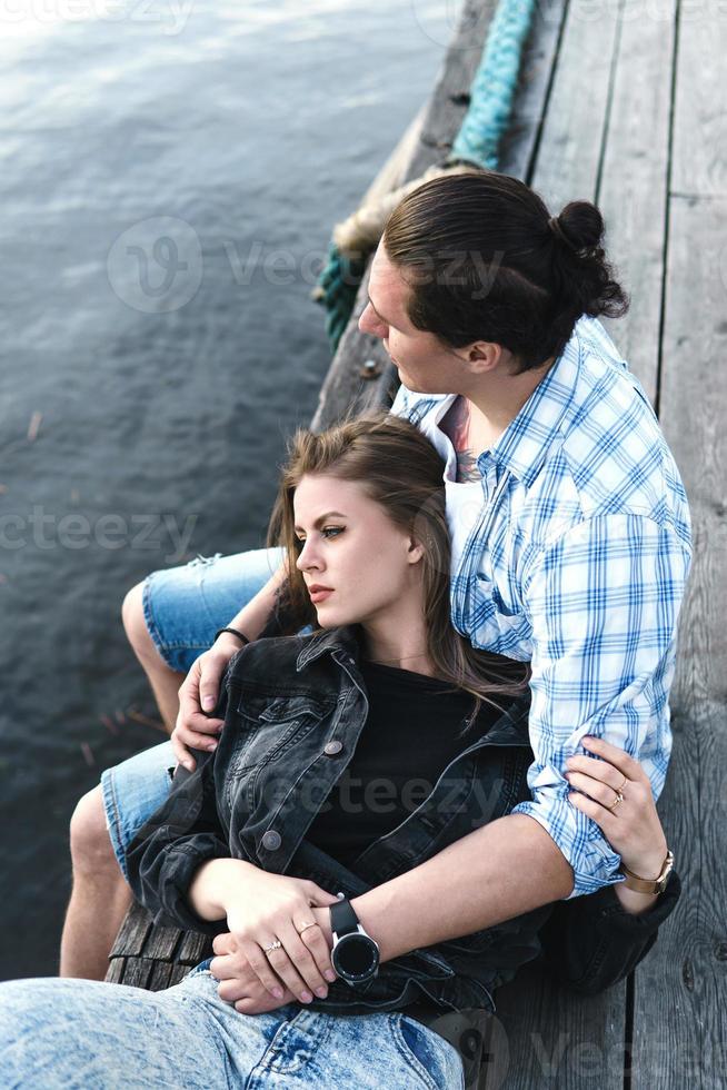 jong sensueel en liefhebbend paar zittend Aan een pier naast een rivier- foto