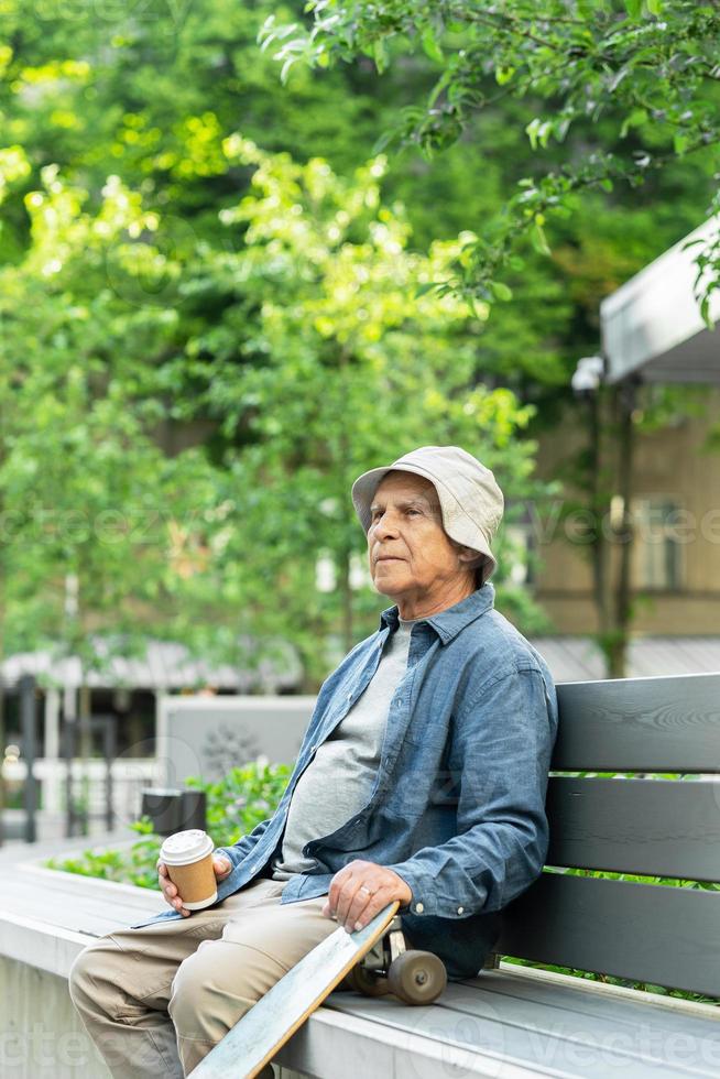 ouderen Mens met een longboard zittend Aan de bank en drinken koffie in een stad park foto