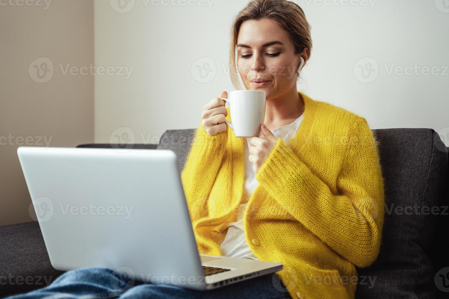 vrouw met kop van heet koffie zittend Aan de sofa en gebruik makend van laptop computer Bij huis foto