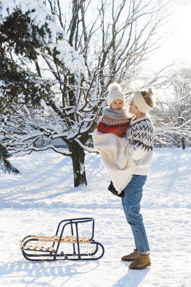 jong moeder en haar schattig weinig zoon met retro slee in een besneeuwd park gedurende zonnig dag foto