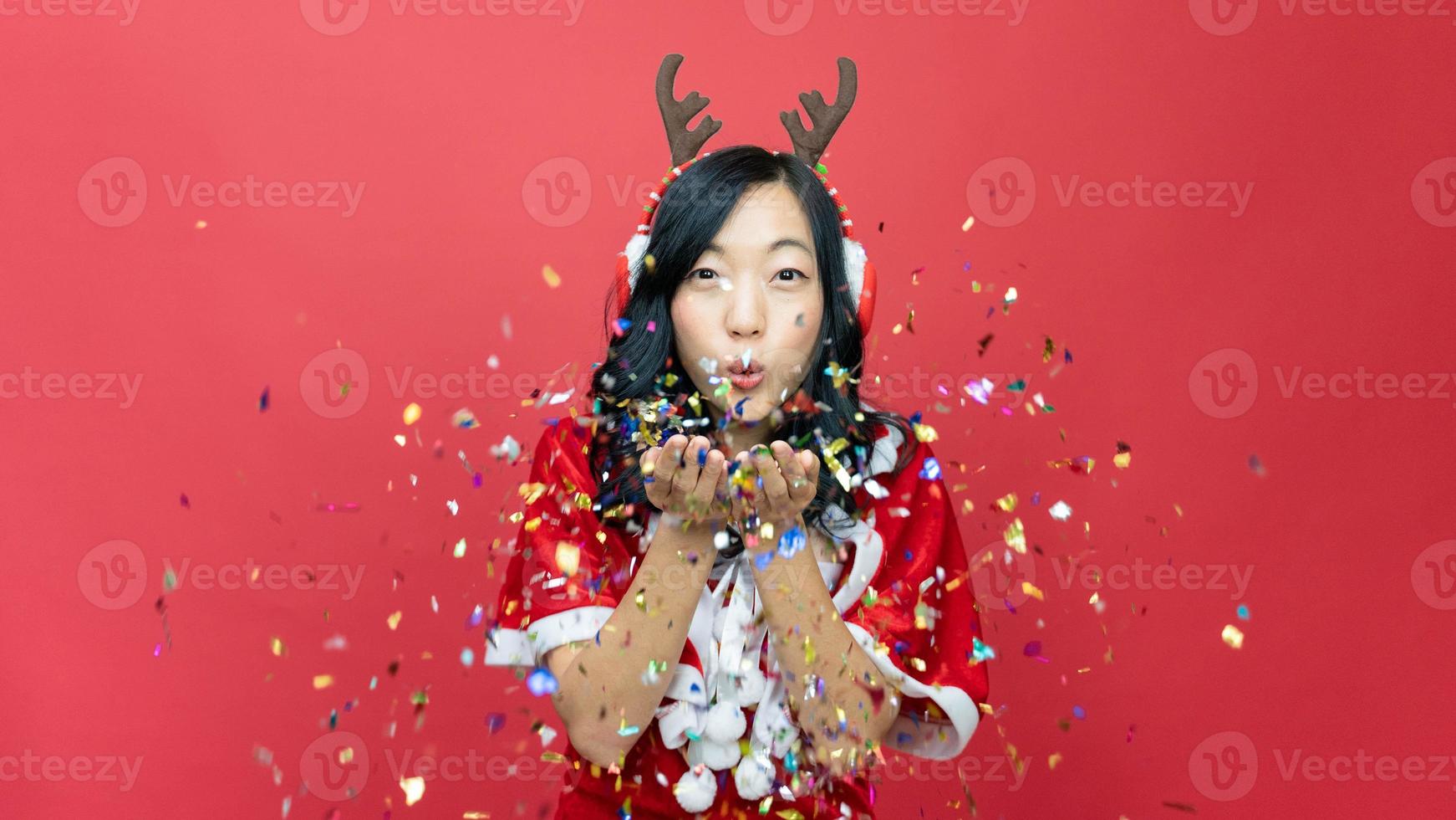 gelukkig lief vrouw met rood santy kostuum Bezig met verzenden een kus naar camera Aan rood achtergrond met confetti. concept van de nieuw jaar en Kerstmis dag. foto