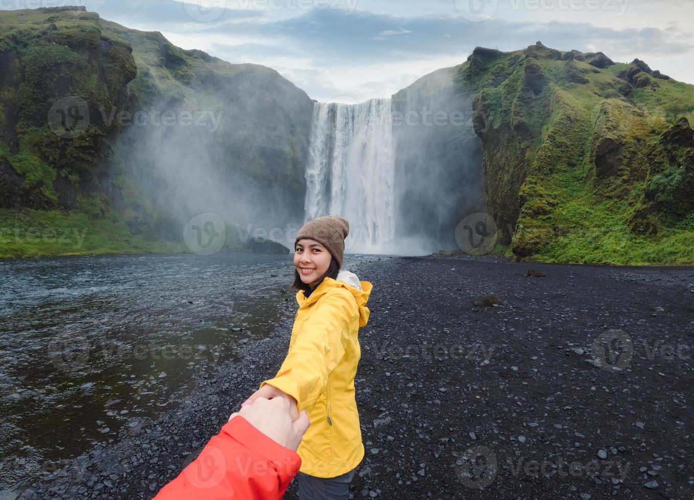 Aziatisch vrouw Holding handen met paar Bij voorkant van skogafoss waterval vloeiende Aan klif in zomer Bij IJsland foto