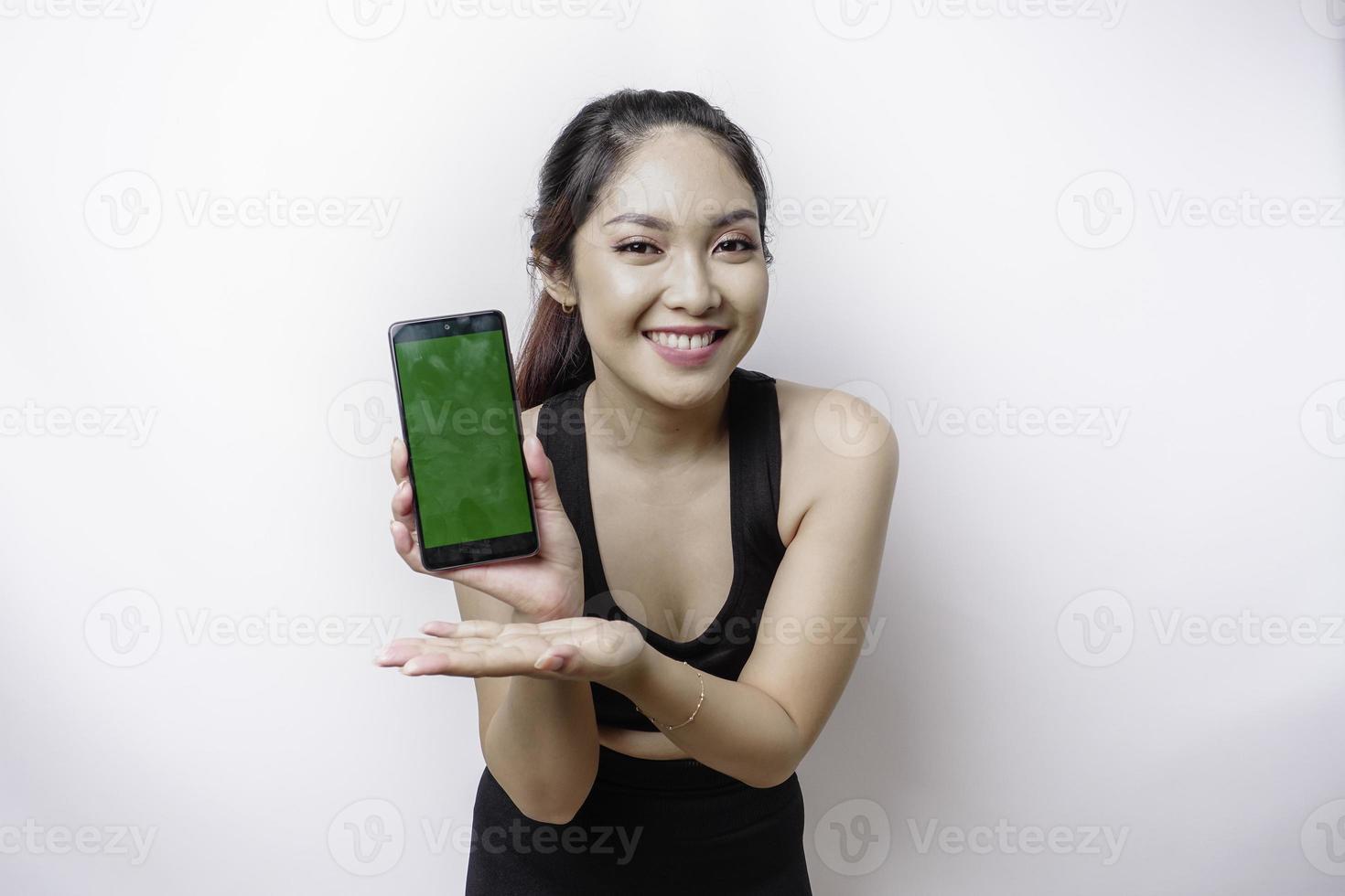 een portret van een sportief Aziatisch vrouw vervelend een sportkleding en tonen groen scherm Aan haar telefoon, geïsoleerd door wit achtergrond foto