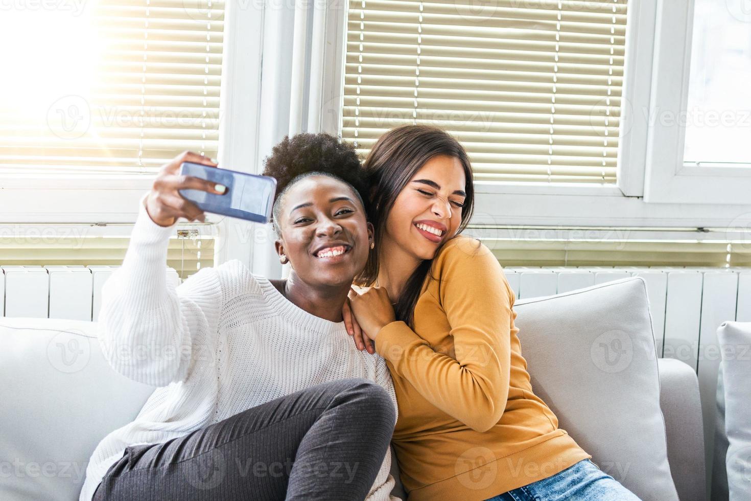 lachend vrouw vrienden knuffelen elk andere Aan sofa terwijl nemen selfie foto Aan slim telefoon. lief Kaukasisch en Afrikaanse Amerikaans meisjes uitdrukken positief emoties naar camera.