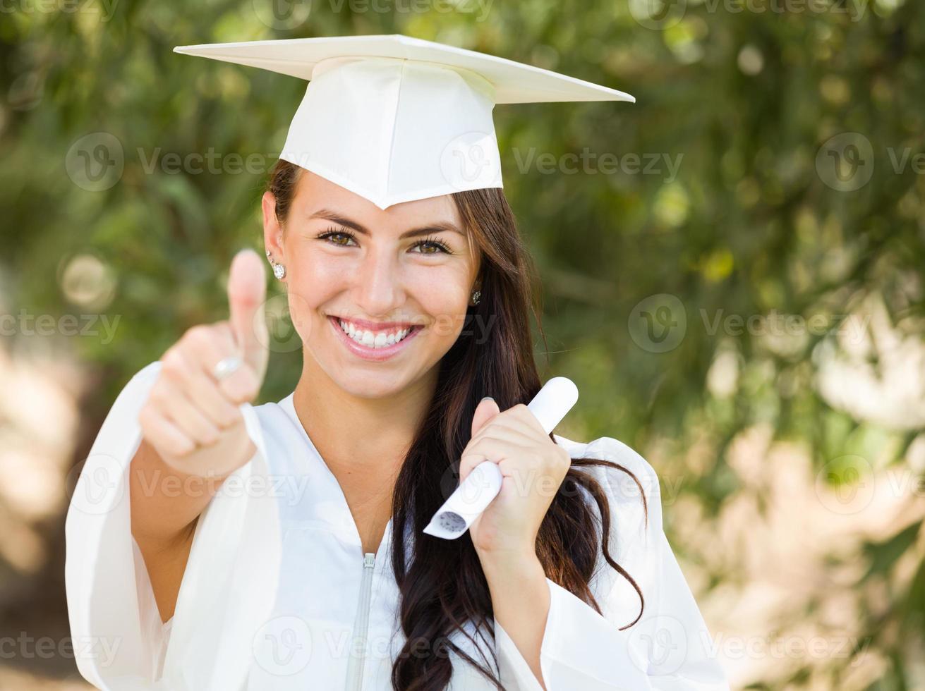 gemengd ras duimen omhoog meisje vieren diploma uitreiking buiten in pet en japon met diploma in hand- foto