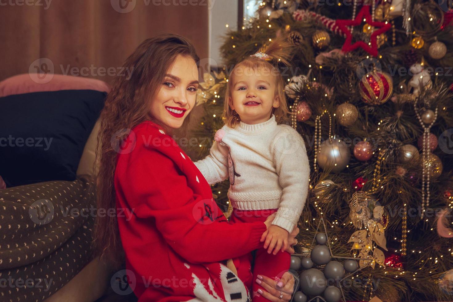 jong vrolijk mam knuffels haar weinig baby meisje Bij de Kerstmis boom foto