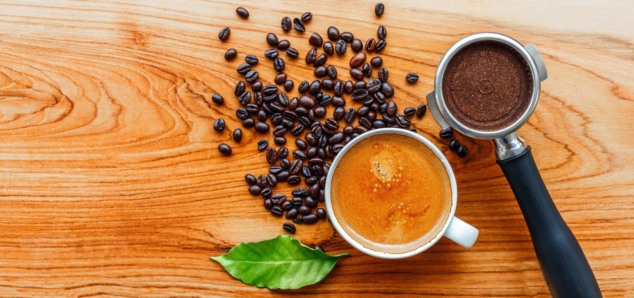 top visie van espresso koffie kop en uitrusting van barista koffie gereedschap filterhouder en donker geroosterd koffie bonen met groen koffie blad Aan houten tafel foto