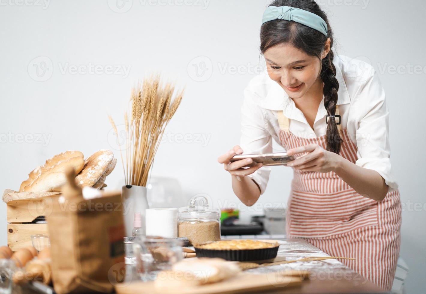 jong vrouw draagt schort nemen selfie foto met eigengemaakt taart in keuken. portret van mooi Aziatisch vrouw bakken toetje en hebben pret nemen foto door smartphone voor online sociaal. huis koken.