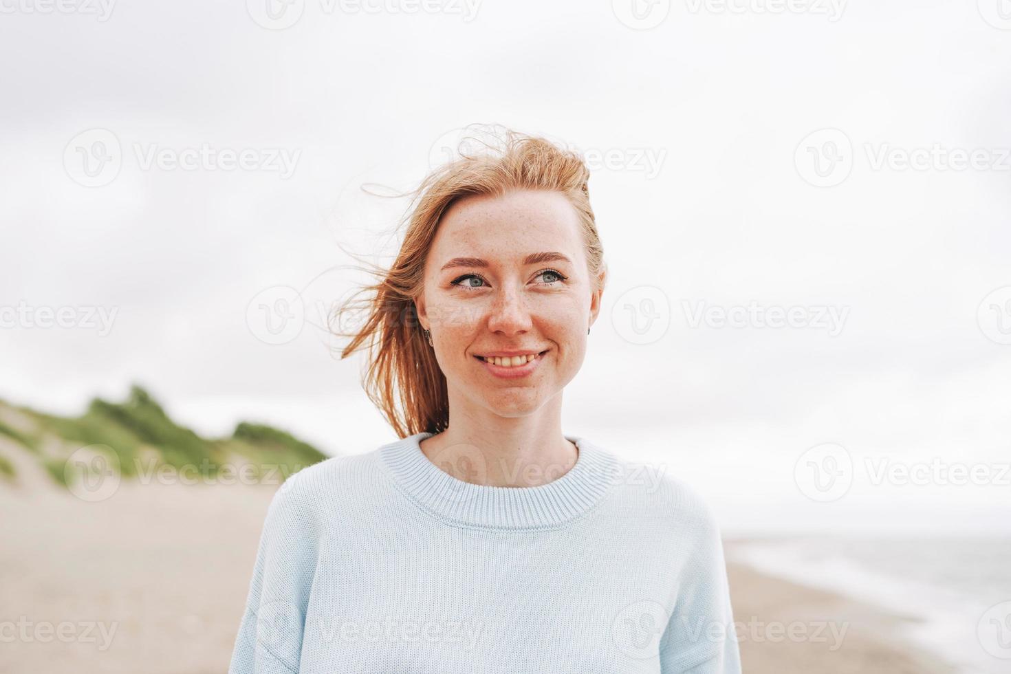 portret van jong rood haren vrouw in licht blauw trui Aan zand strand door zee in storm foto