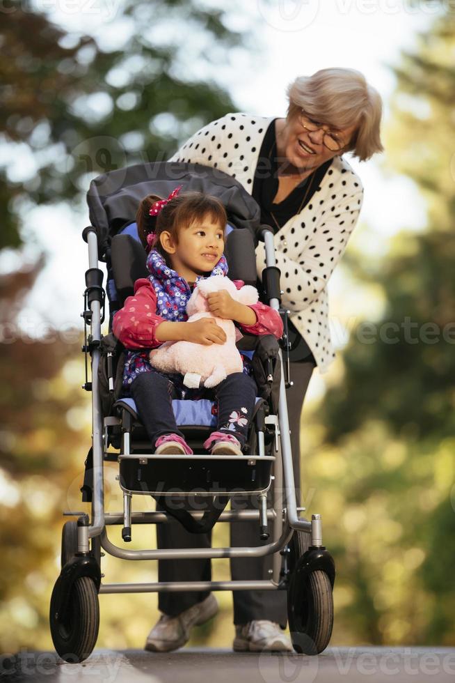gelukkig senior dame voortvarend wiel stoel en kinderen. grootmoeder en kinderen genieten van een wandelen in de park. kind ondersteunen gehandicapt grootouder. familie op bezoek komen. generaties liefde en verhouding foto
