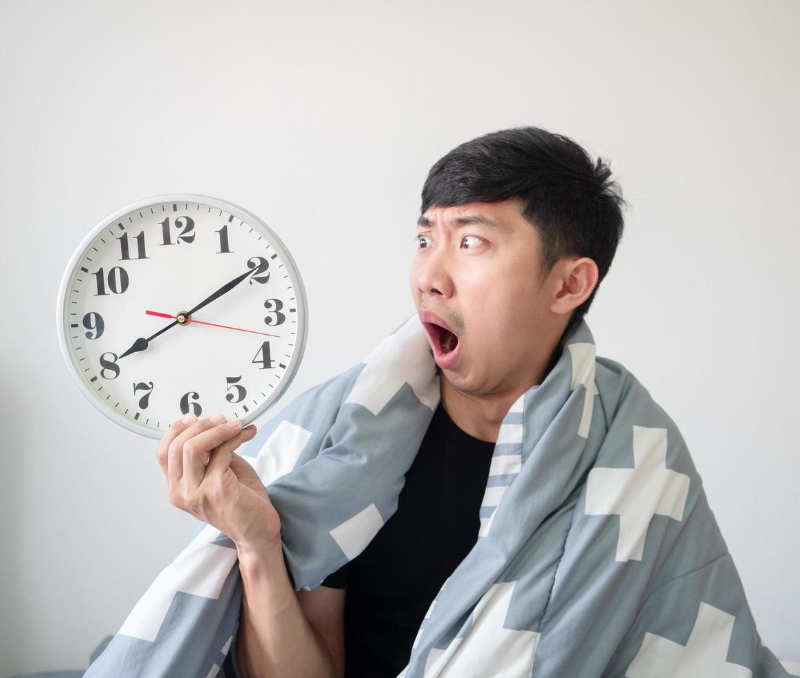 Aziatisch Mens met deken Hoes lichaam wakker worden omhoog laat voelen geschokt en kijken Bij klok in hand- Aan wit geïsoleerd foto