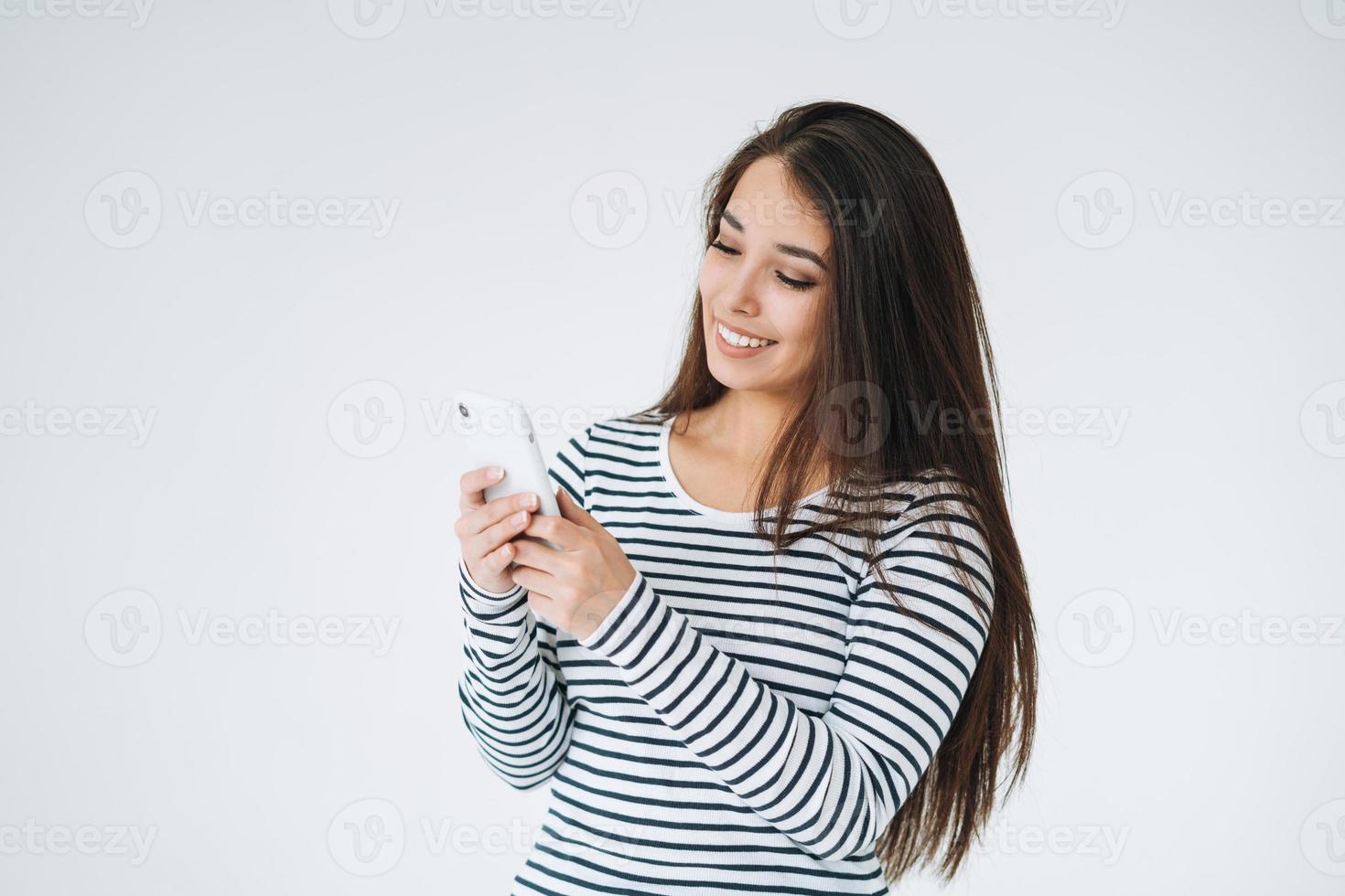 jong gelukkig Aziatisch vrouw met lang haar- in gestreept lange mouw gebruik makend van mobiel telefoon Aan wit achtergrond geïsoleerd foto