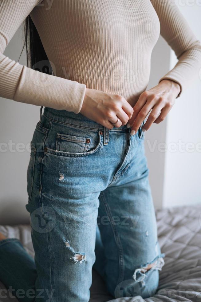 Bijsnijden foto van jong glimlachen vrouw tiener meisje met donker lang haar- in jeans zittend Aan bed Bij huis