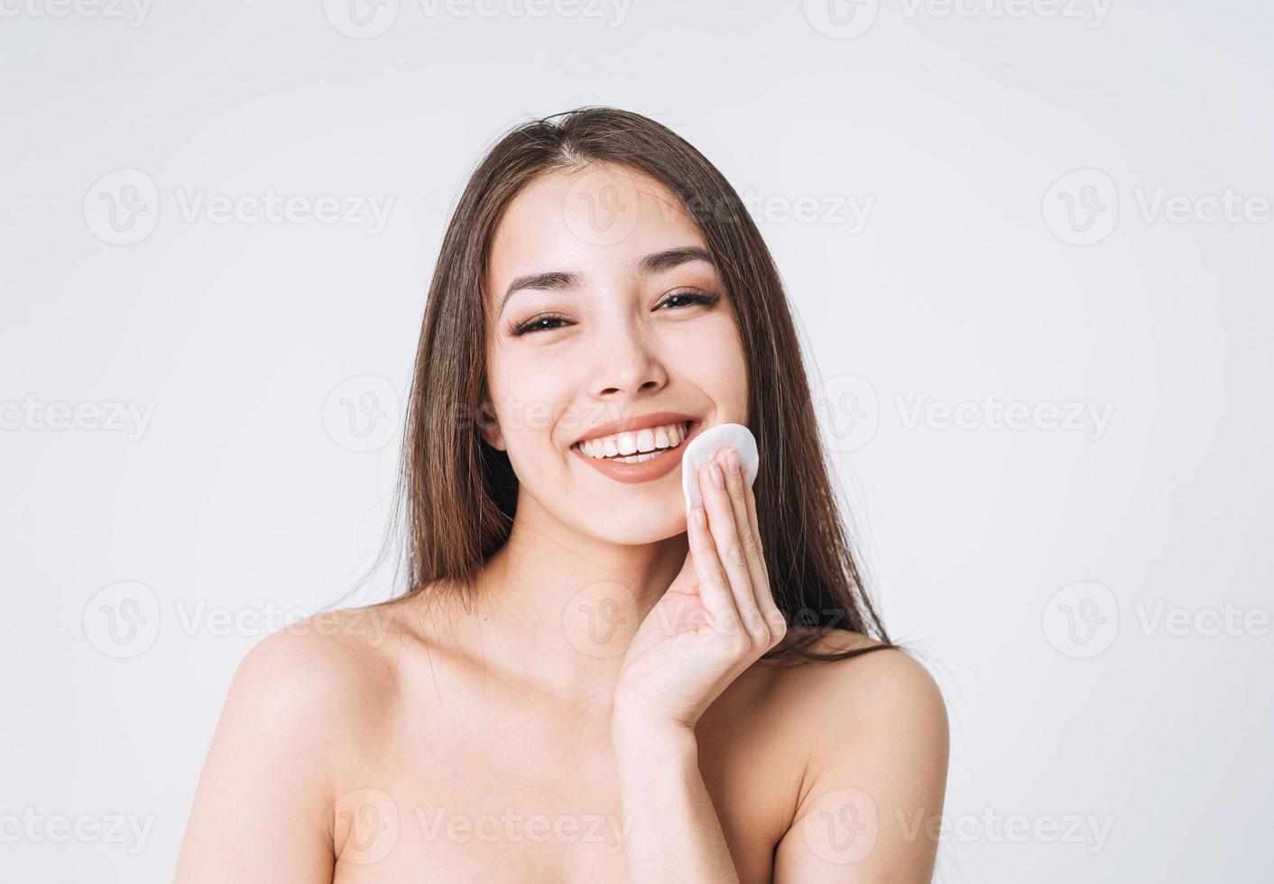 schoonheid portret van gelukkig glimlachen Aziatisch vrouw met donker lang haar- met katoen stootkussen Aan schoon vers huid gezicht en handen Aan wit achtergrond geïsoleerd, huid reiniging en Verwijderen bedenken foto