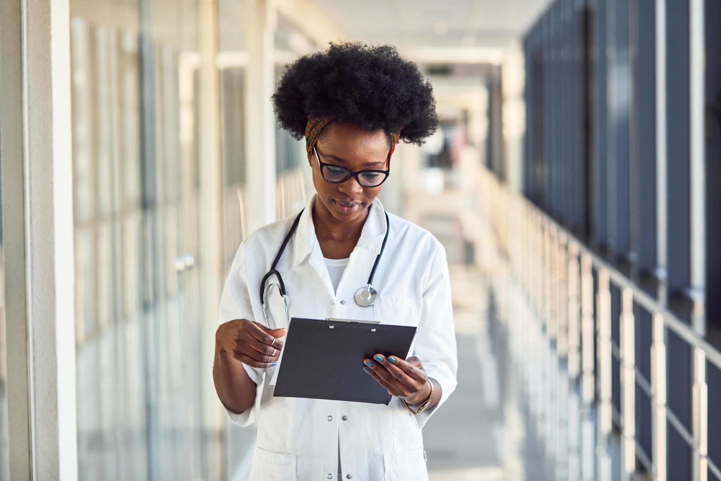 jong Afrikaanse Amerikaans vrouw dokter in wit uniform met stethoscoop en kladblok staand in gang foto