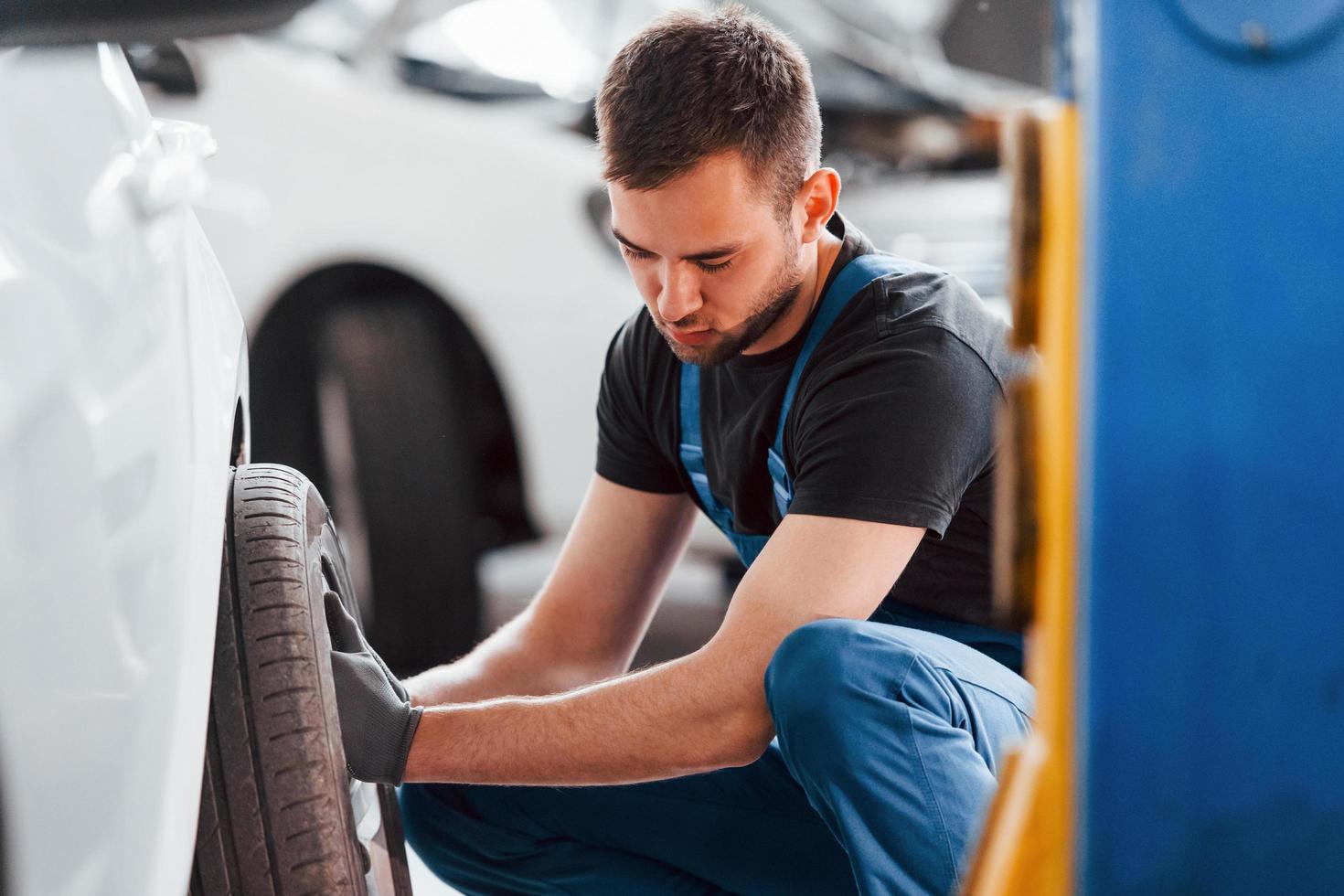 Mens in werk uniform zittend en veranderen auto wiel binnenshuis. opvatting van auto- onderhoud foto