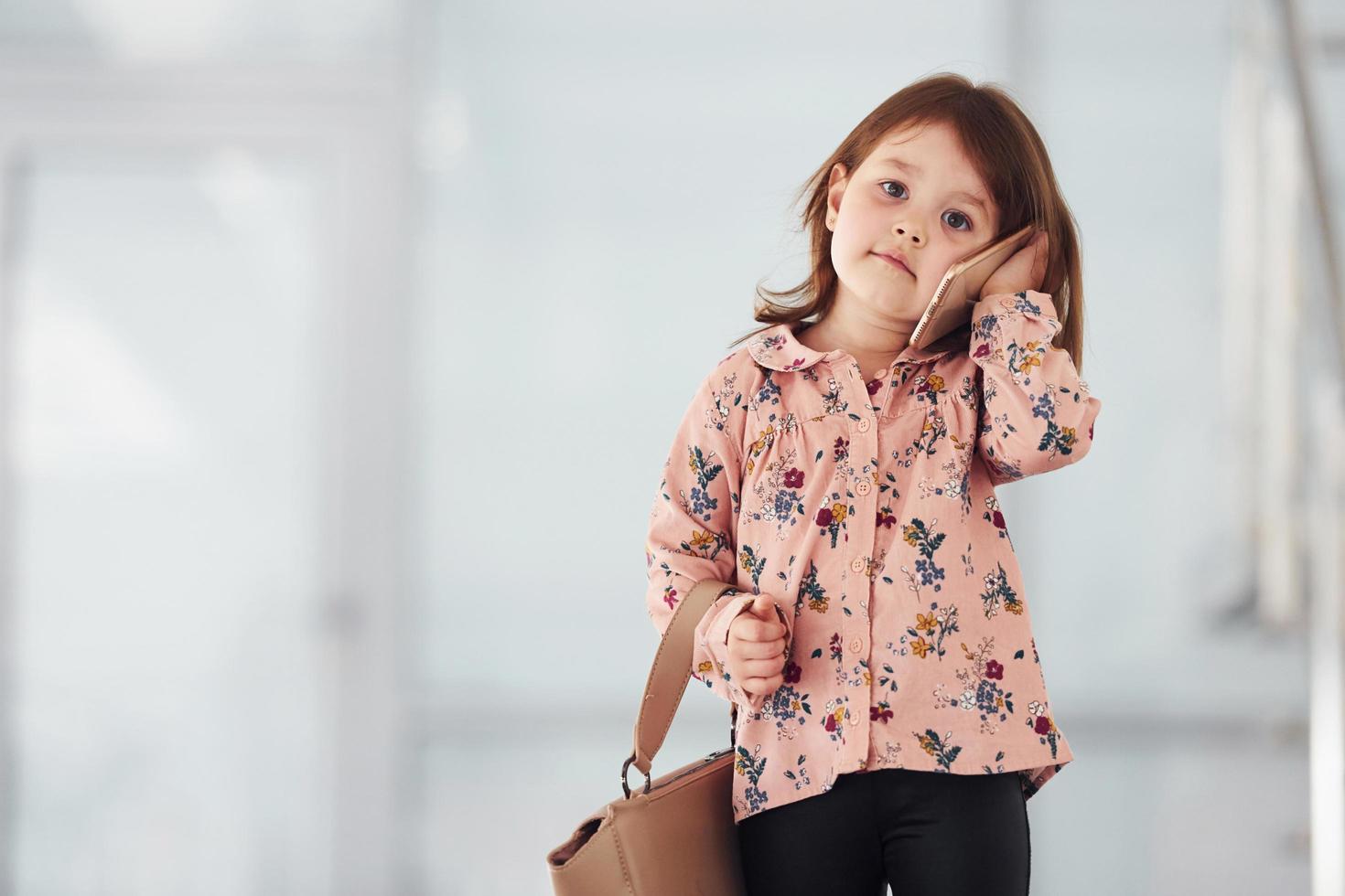 schattig weinig meisje met zak binnenshuis in de kantoor of luchthaven pratend door telefoon foto