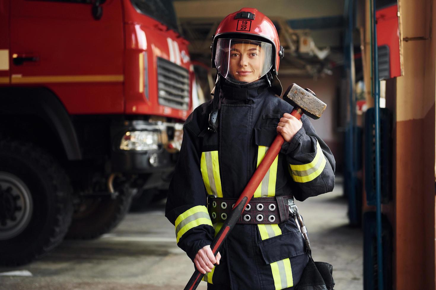 met hamer in handen. vrouw brandweerman in beschermend uniform staand in de buurt vrachtauto foto