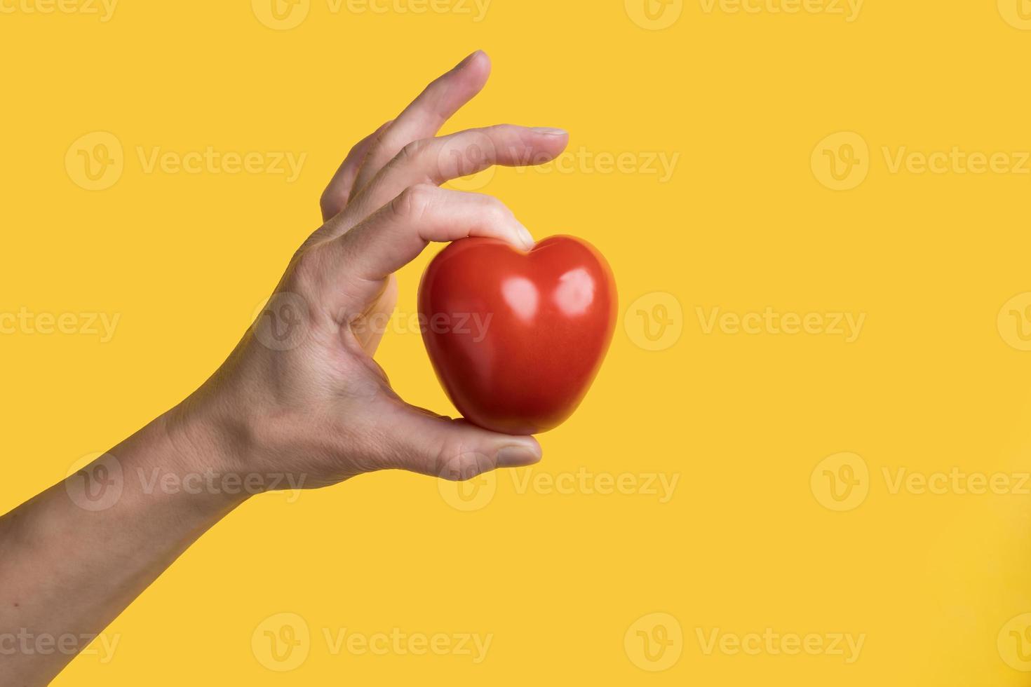 hand- houdt een rood tomaat in de het formulier van een hart, een symbool van liefde, Aan een geel achtergrond. gezond voedsel concept. Valentijnsdag dag. foto